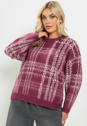Femme aux cheveux blonds portant un pull à carreaux rose et violet en matière duveteuse et un pantalon gris sur un fond blanc uni.