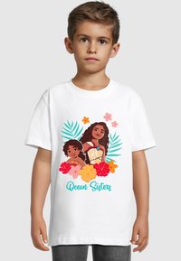 Camiseta blanca con un diseño impreso colorido de dos chicas con acentos florales, rodeadas de hojas tropicales y el texto "Hermanas del Océano".