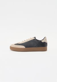 Sko med svart lær overdel, beige semsket tåhette og brun gummisåle. Har flate lisser og preget merkevarelogo på hælen.
