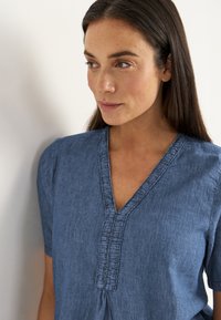 Frau mit langen, dunklen Haaren, die ein blaues V-Ausschnitt-Oberteil mit dekorativen Nähten trägt und leicht zur Seite schaut, vor einem schlichten Hintergrund.