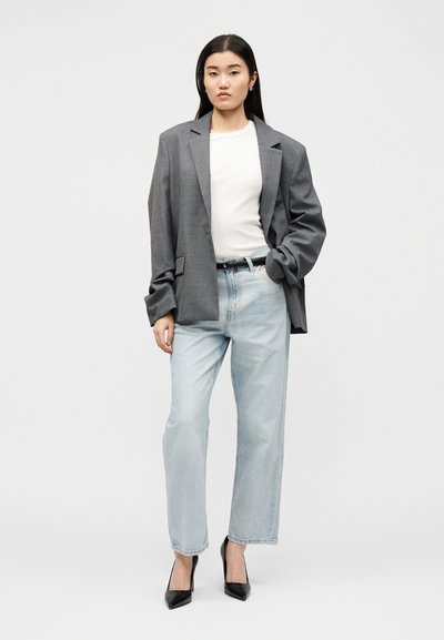 Νεαρή γυναίκα που φοράει γκρι oversized σακάκι, λευκό τοπ, ανοιχτό μπλε τζιν, μαύρη ζώνη και μαύρες ψηλοτάκουνες γόβες, στέκεται μπροστά σε ένα απλό φόντο.