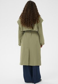 Olivno zeleni trenč kot z pasom, širokimi rokavi in hrbtnim režo, v kombinaciji z širokimi jeans hlačami in vidnimi kodrastimi lasmi.