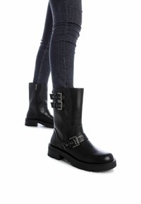 Botas negras de cuero hasta el tobillo con suela gruesa, que cuentan con múltiples detalles de hebillas y un zipper lateral. Llevadas con jeans oscuros.