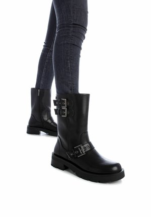 Botas negras de cuero hasta el tobillo con suela gruesa, que cuentan con múltiples detalles de hebillas y un zipper lateral. Llevadas con jeans oscuros.