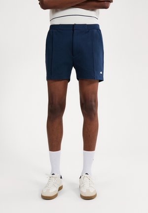 Mann trägt maßgeschneiderte marineblaue Shorts, weiße Socken und weiße Sneakers, steht mit verschränkten Armen vor einem schlichten Hintergrund.
