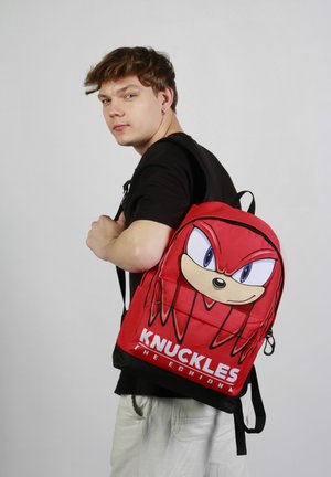 Sac à dos rouge avec un grand visage de Knuckles en dessin animé, accents noirs, sangles ajustables et un compartiment zippé sur le côté.