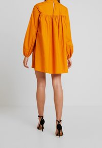 Lyhyt oranssi mekko, jossa on ilmapallohihat, poimutettu selkäpuoli ja napitus kaulassa, yhdistetty mustiin korkokenkiin.