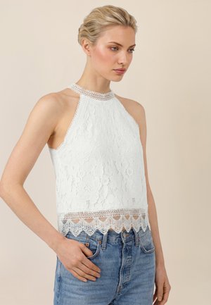 Young woman with blonde hair wearing a sleeveless white lace halter top and blue jeans, standing against a beige background.