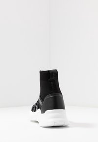 Černé vysoké tenisky s hladkým syntetickým materiálem a bílou podrážkou. Mají pletený svršek a akcenty loga na boku a patě.