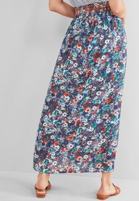Femme portant une maxi-jupe florale colorée avec une taille élastique et des sandales à enfiler marron sur un fond gris.