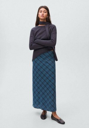 Pull en tricot gris foncé avec un design enveloppant, associé à une longue jupe en tartan bleu avec des lignes plus foncées, et des chaussures noires plates avec desclous.