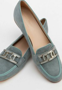 Blaue Wildleder-Loafer mit einem metallischen Kettenakzent, abgerundetem Zehenbereich, kontrastierendem hellblauen Rand und einer glatten beige Innensohle.