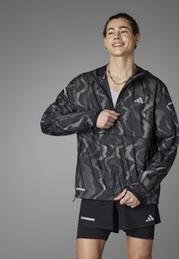Schwarze Jacke mit einem kammoskopischen Muster aus weißen Punkten und Reißverschluss, getragen über einem weißen Tanktop, kombiniert mit schwarzen Shorts mit Akzenten.