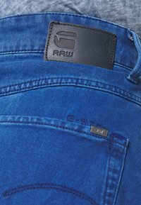 Blå denimjeans med en svart läderpatch från G-Star RAW på midjebandet och synlig sömnad på bakfickan.