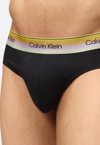 Άνδρας που φοράει μαύρα εσώρουχα με ζώνη μέσης σε αποχρώσεις κίτρινου-μωβ με το λογότυπο Calvin Klein σε μαύρο κείμενο.