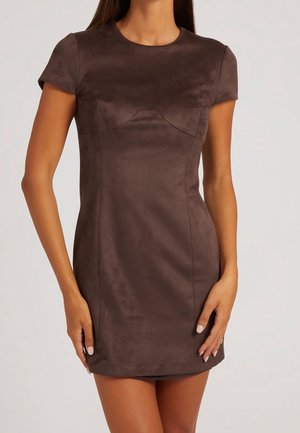 Femme portant une mini-robe ajustée à manches courtes en suède marron foncé, debout devant un fond clair uni.