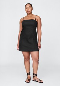 Čierne minišaty bez rukávov s tenkými ramienkami a priliehavým živôtikom. Jednoduchý dizajn, hladká textúra. Nositý s čiernymi sandálmi na remienky.