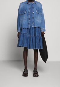 Denimová bunda se dvěma náprsními kapsami a knoflíky, spárovaná s vrstvenými denimovými šaty. Černé ploché sandály dokončují vzhled.