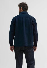 Námořnická modrá svrchní bunda s dlouhými rukávy a límcem. Vyrobena z pevného materiálu, má volný střih a knoflík na manžetě.