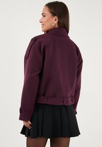 Vijolična jakna iz volnenega mešanice z epauleti na ramenskem delu, zapenjanjem v pasu in gumbnimi manšetami, v kombinaciji s črno nabirano krilo.