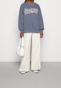 Blå sweatshirt med texten "HOLLISTER CALIFORNIA", tillsammans med vita vida byxor och en liten vit väska. Slätt tyg, avslappnad passform.