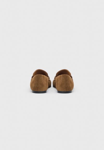 Ζευγάρι καφέ σαουέτ μοκασίνια χωρίς κορδόνια, εμφανίζονται από πίσω σε απλό φωτεινό φόντο.