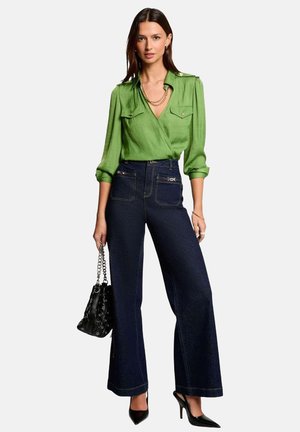 Blouse en soie verte avec deux poches avant, associée à un pantalon en denim foncé à taille haute et évasé. Le mannequin tient un sac à main noir et porte des talons noirs.