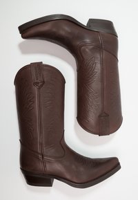 Las botas de vaquero de cuero marrón tienen una punta afilada, tacón bajo y detalles en relieve en el vástago. El diseño incluye una etiqueta de marca.