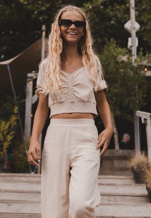Beige cropped top met geplooide details en korte mouwen, gecombineerd met beige wijde broek. Zonnebril maakt de outfit compleet.