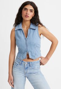 Mulher com cabelo longo e escuro, vestindo um colete sem mangas de denim azul claro com fecho e jeans de cintura alta azul claro com um padrão subtil.