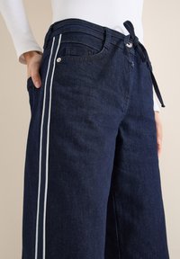 Pantalones de mezclilla azul oscuro de pierna ancha con cintura anudada, con rayas laterales blancas y bolsillos frontales. La textura es suave y ligeramente texturizada.