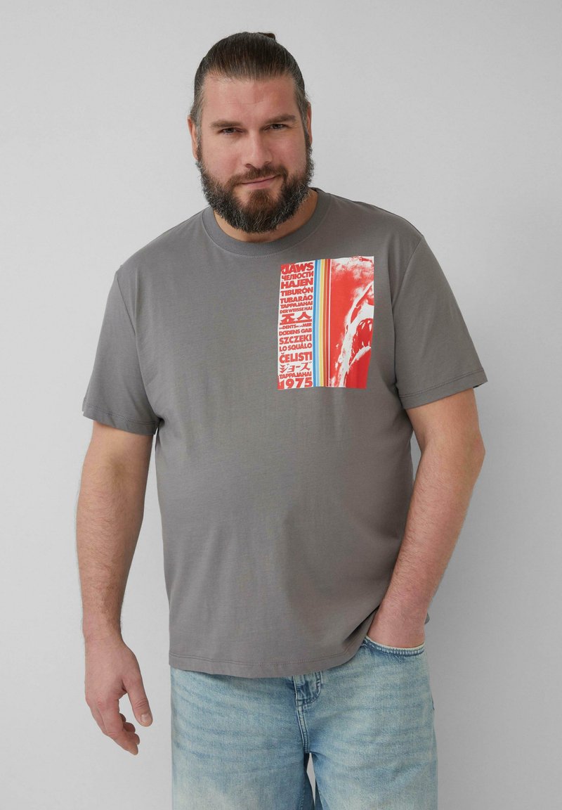 Graues T-Shirt mit kurzen Ärmeln, das einen roten und weißen grafischen Druck mit Text zeigt. Das Shirt hat einen Rundhalsausschnitt und einen lässigen Schnitt. Helle blaue Jeans.