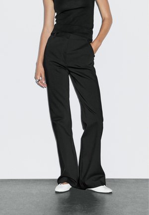 Kvinde iført sorte højtaljede bukser med vide ben, sort ærmeløs top, hvide balletsko, med den ene hånd i lommen og stående på gråt gulv.