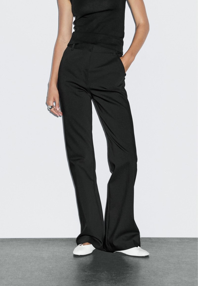 Frau trägt hoch taillierte schwarze Hosen mit weitem Bein, schwarzes ärmelloses Oberteil, weiße Ballettschuhe, steht mit einer Hand in der Tasche auf grauem Boden.