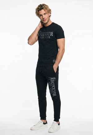 Junger Mann in schwarzem "Boxeur des Rues" T-Shirt und Hose, steht mit einer Hand in der Tasche und der anderen am Hals, trägt weiße Sneakers.