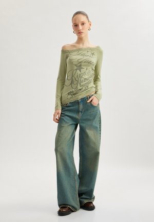Blusa de manga larga tipo off-shoulder en verde claro con estampado gráfico; combinada con jeans de pierna ancha azul, que presentan una textura desgastada y talle alto.