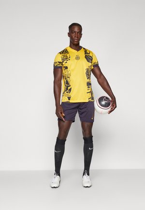 Gul fodboldtrøje med sort grafisk design, parret med mørke shorts, sorte sokker og hvide sko; holder en fodbold.