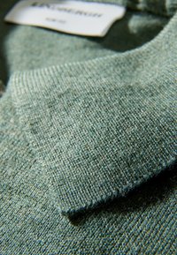 Zielony, teksturowany materiał swetra z wywiniętym kołnierzem i widoczną metką. Splot jest drobny, a powierzchnia miękka i gładka.