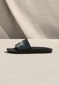 Μαύρο σανδάλι slide με λευκό κείμενο "White" στο λουρί, τοποθετημένο σε ανοιχτόχρωμη επιφάνεια με φόντο μπεζ ύφασμα.