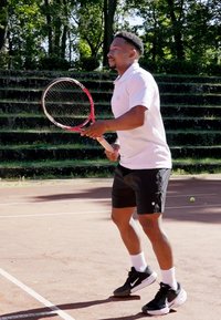 Jogador de ténis com uma camisola polo branca e calções pretos segura uma raquete de ténis com detalhes vermelhos num campo de terra batida rodeado de vegetação.
