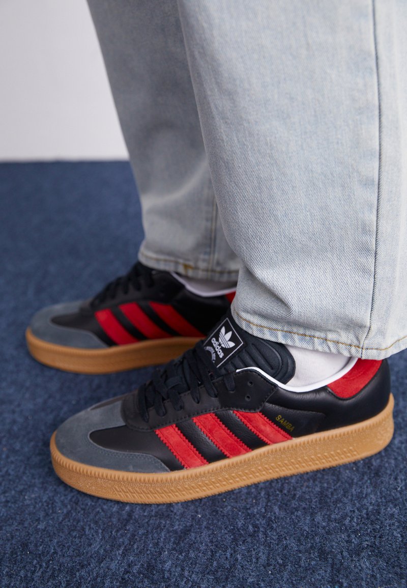Czarne i szare sneakersy z czerwonymi paskami, teksturowana cholewka i gumowa podeszwa. Posiadają logo na języku oraz zamszowy detal na palcach.