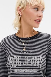 Pruhovaná sivá košeľa s dlhými rukávmi s potlačou "BDG JEANS", okrúhlym výstrihom a vrstvenými zlatými náhrdelníkmi s vzorovanými príveskami.