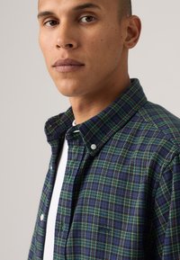 Flannel shirt in dark blue and green with a checkered pattern. Features button front, collar, and white undershirt visible beneath.