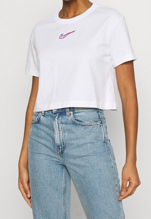 Persoon die een witte cropped T-shirt draagt met een paars Nike-logo en lichtblauwe denim jeans met hoge taille, staand tegen een effen achtergrond.
