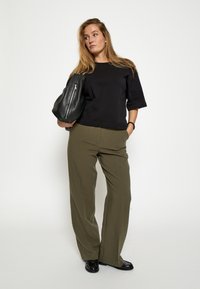 Zwarte, mouwloze top met een ronde hals, gecombineerd met hoge, olijfgroene broeken. Model houdt een zwarte tas vast en staat tegen een effen achtergrond.