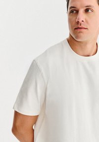 Młody mężczyzna stoi zwrócony w prawo, ubrany w gładką, białą koszulkę z krótkim rękawem na jasnym, neutralnym tle.
