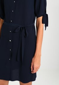Námořnická modrá šaty vyrobené z lehkého materiálu, s knoflíkovou přední stranou a stříbrným kováním a s vázáním v pase pro nastavitelné padnutí. Krátké rukávy s vázáním.