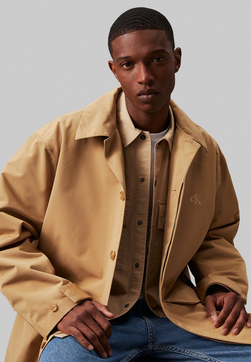 Beżowy wodoodporny płaszcz przeciwdeszczowy z dużym kołnierzem, zapięciem na guziki i detalem z logo. Nosi go na jasnobrązowej koszuli i dżinsowych spodniach.