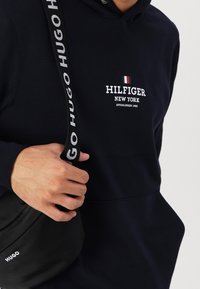 Sudadera negra con el logo "HILFIGER" en blanco y un acento en rojo y azul. Se lleva con un bolso cruzado negro que presenta el logo "HUGO".