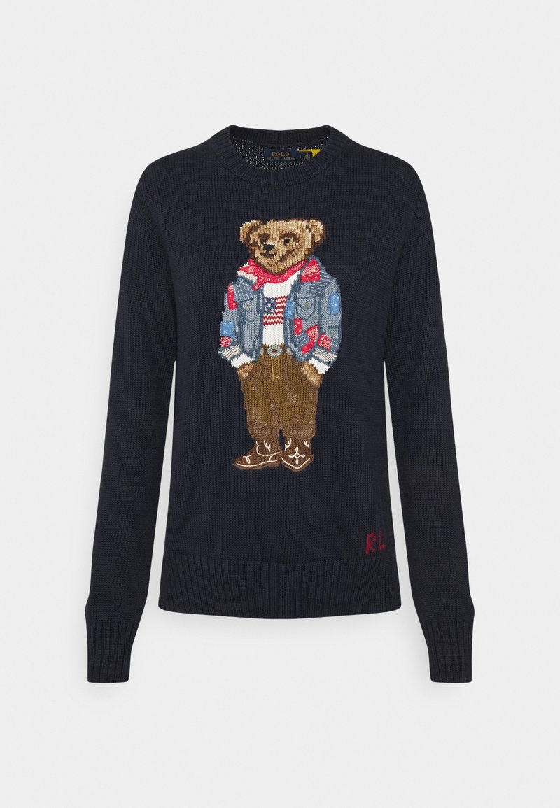 Pull en maille bleu marine avec un ours brodé portant une veste en jean, un bandana rouge, un pull blanc avec un drapeau, un pantalon marron et des bottes.